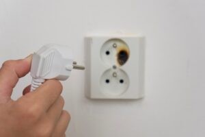 Residential electricians Fusion Electric Kansas City AdobeStock_1667188344 A hand holding an electrical plug near a burnt out and damaged socket, concept of business electrical services, fire safety, home repair.